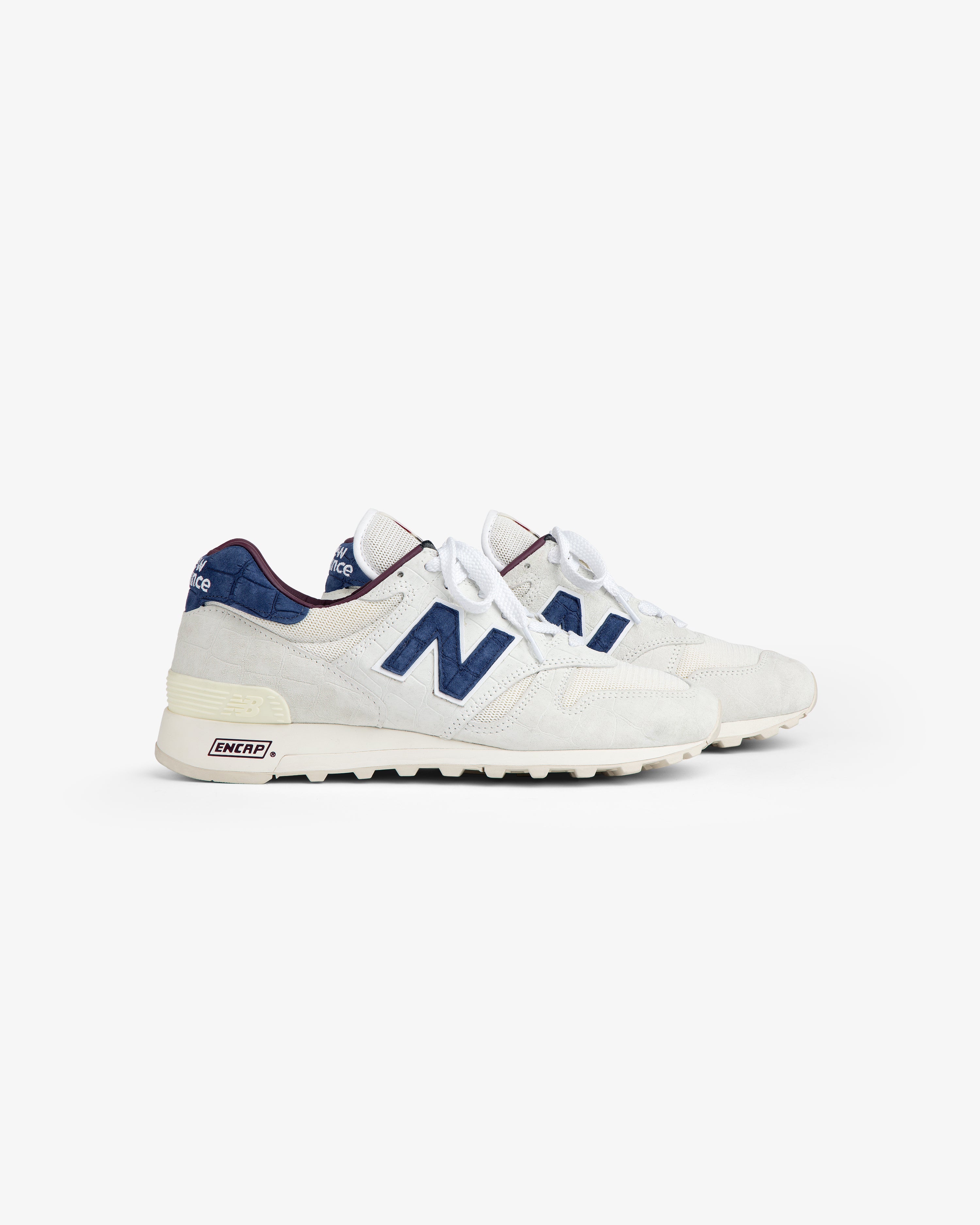 New Balance Made in USA 1300: white sneakers with navy blue N logos, blue accents, and white laces on white background.