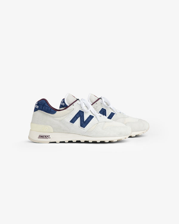 New Balance Made in USA 1300: white sneakers with navy blue N logos, blue accents, and white laces on white background.