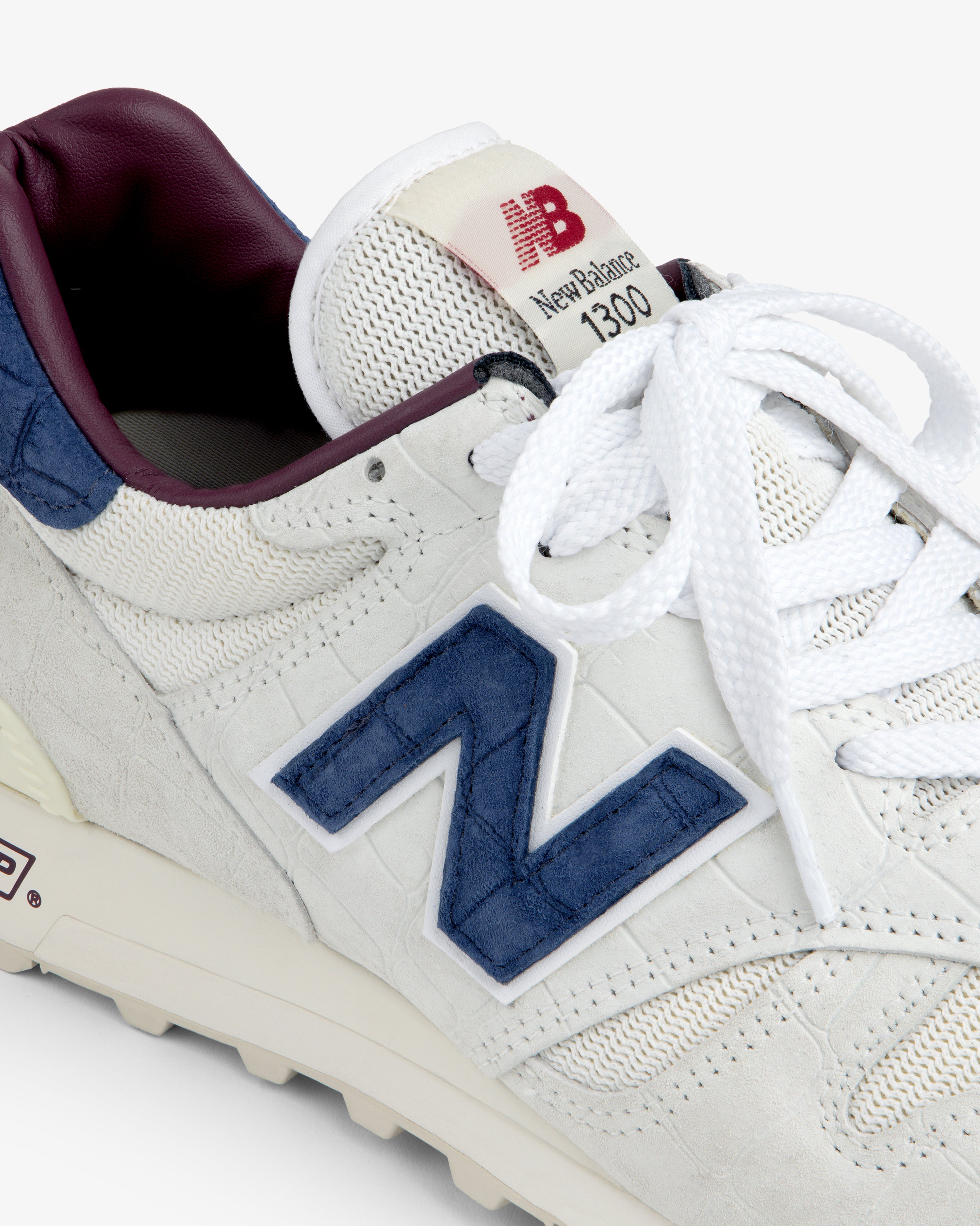 Close-up of a white New Balance Made in USA 1300 with blue logo, maroon lining, and white laces.