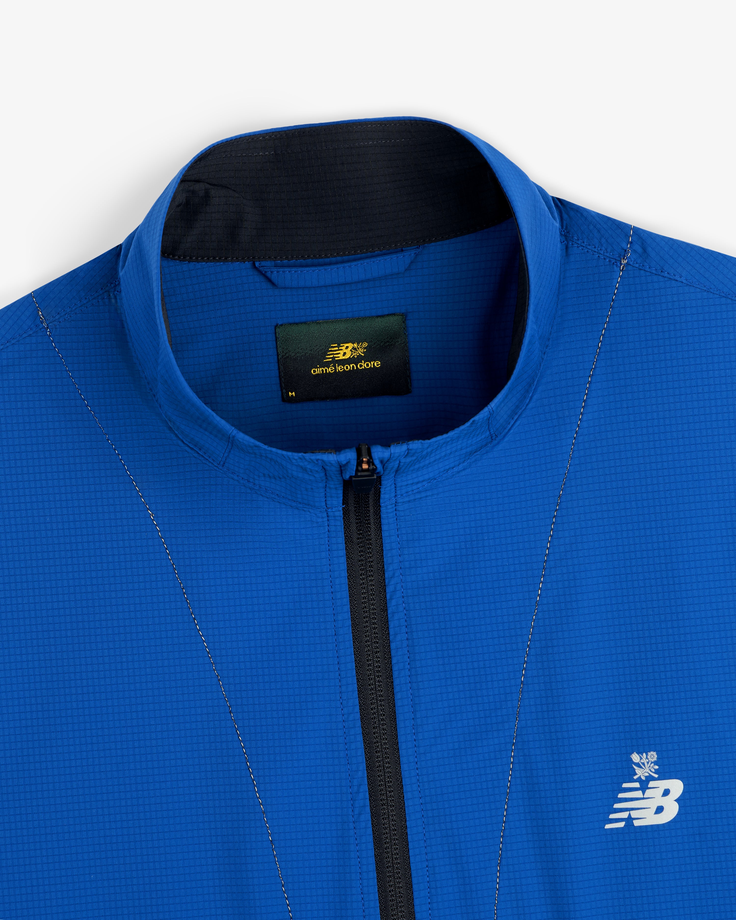 Close-up of the ALD / New Balance Performance Nylon Jacket in blue with a zipper and white embroidered logo near the collar.