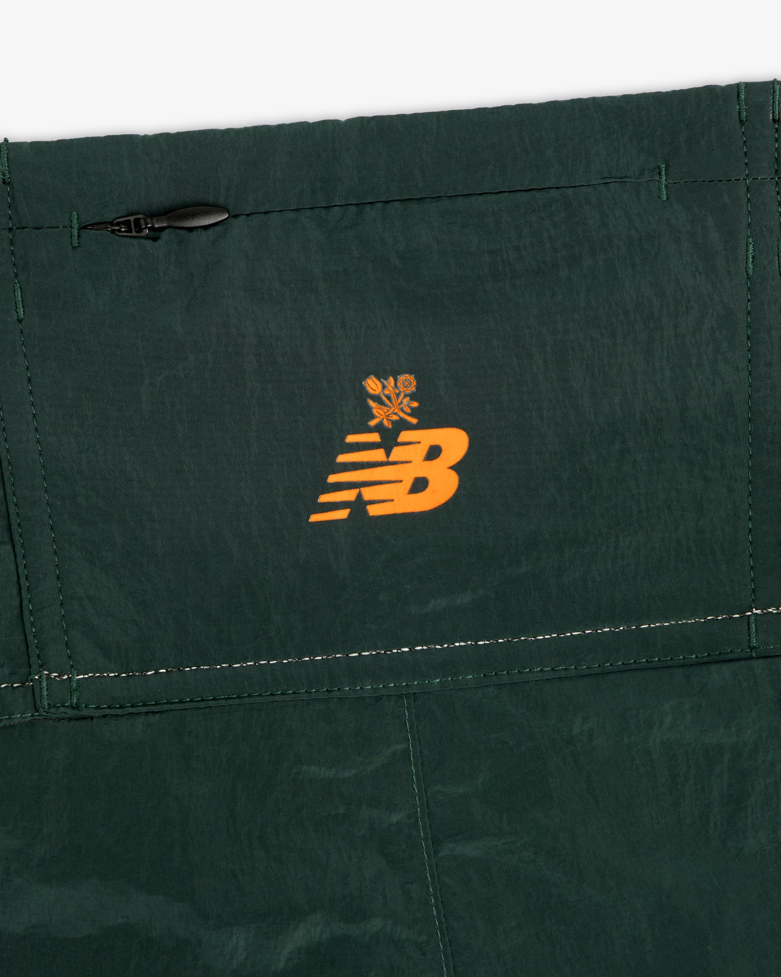 Close-up of dark green ALD / New Balance Lightweight Nylon Warmup Pant with orange logo and flower graphic above seams.