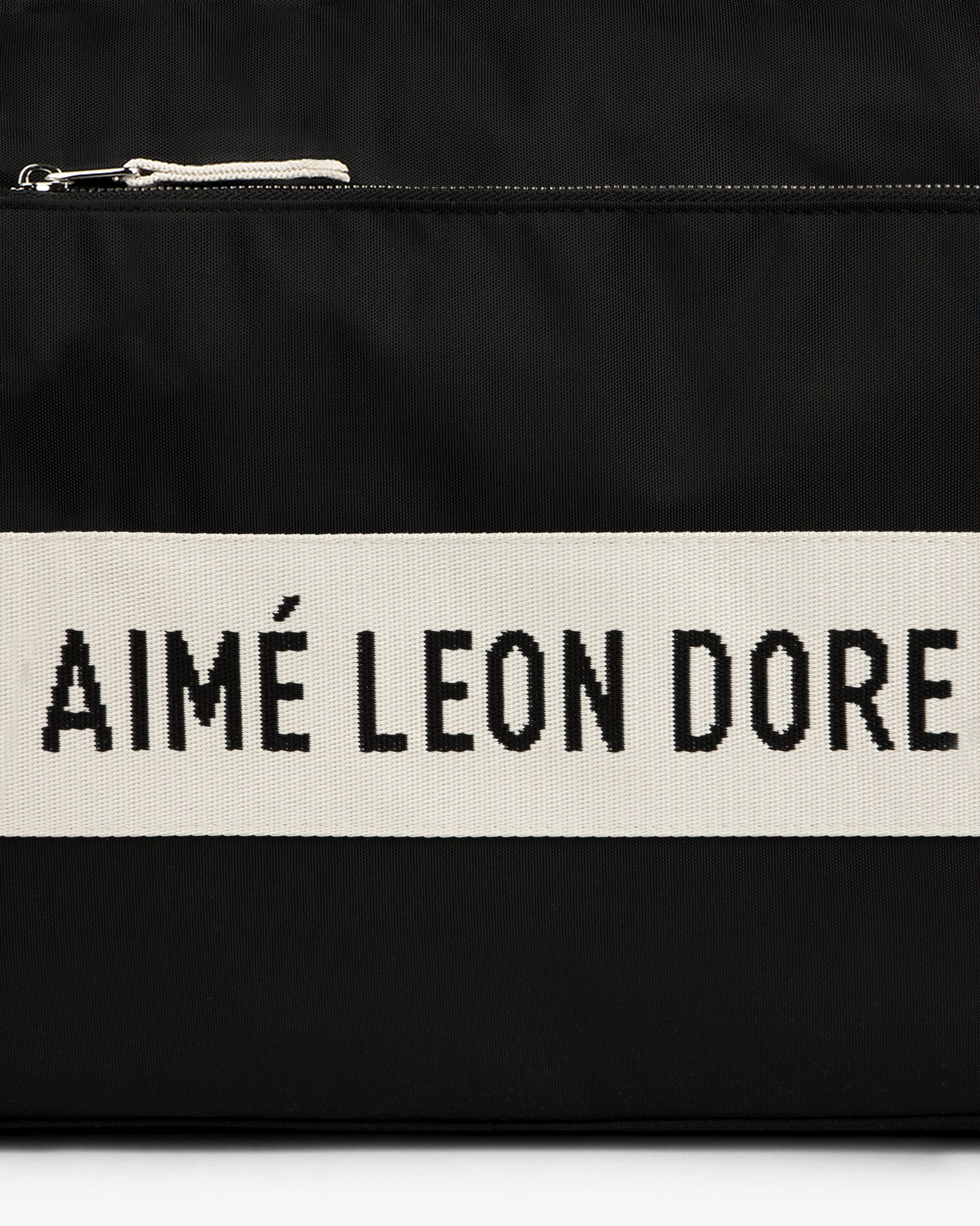 Black nylon duffle bag with a white stripe featuring "AIMÉ LEON DORE" text in bold black letters.