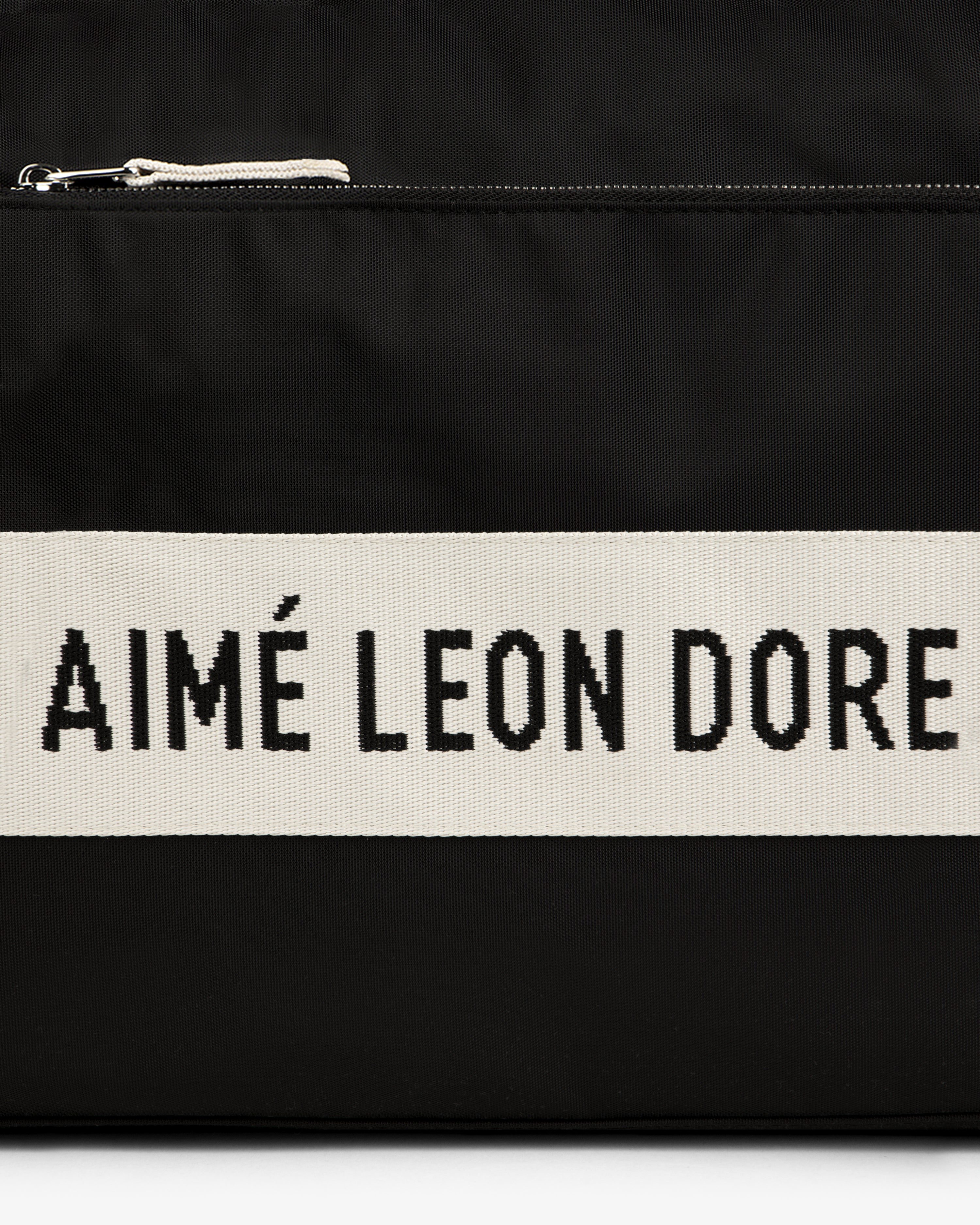 Black nylon duffle bag with a white stripe featuring "AIMÉ LEON DORE" text in bold black letters.