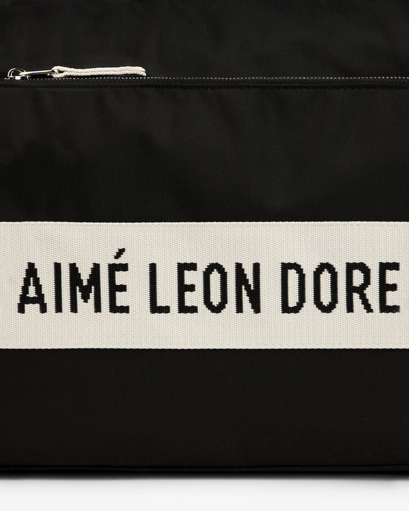 Black nylon duffle bag with a white stripe featuring "AIMÉ LEON DORE" text in bold black letters.