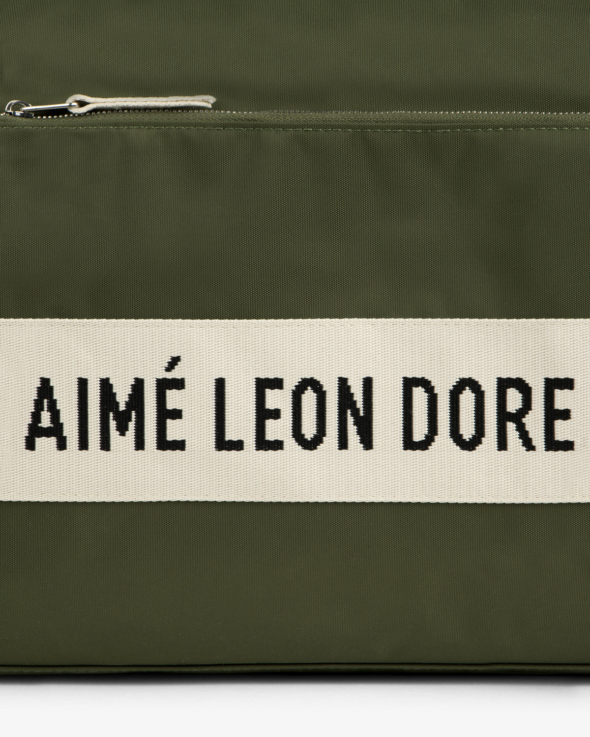 Close-up of an olive green Nylon Duffle Bag with a zipper and a white strip reading AIMÉ LEON DORE in black text.