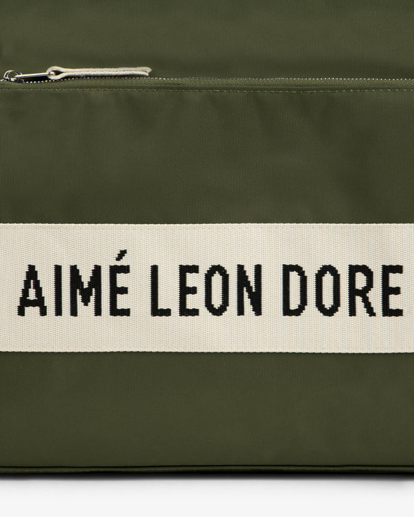 Close-up of an olive green Nylon Duffle Bag with a zipper and a white strip reading AIMÉ LEON DORE in black text.