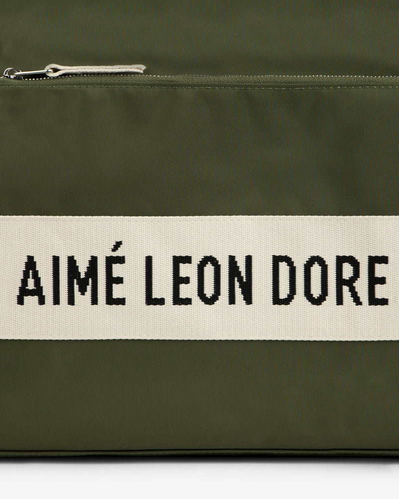 Close-up of an olive green Nylon Duffle Bag with a zipper and a white strip reading AIMÉ LEON DORE in black text.