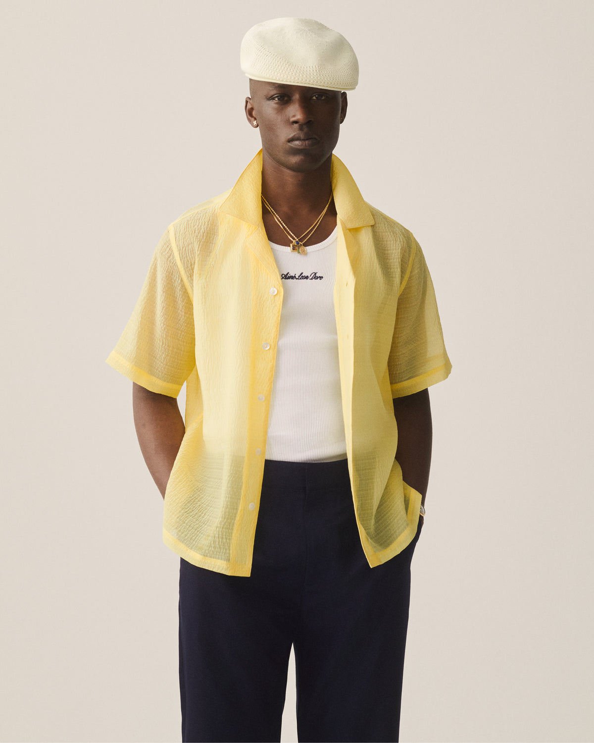 Man sporting an Embroidered Logo Tank Top under a yellow shirt, dark pants, layered necklaces, and a white beret.