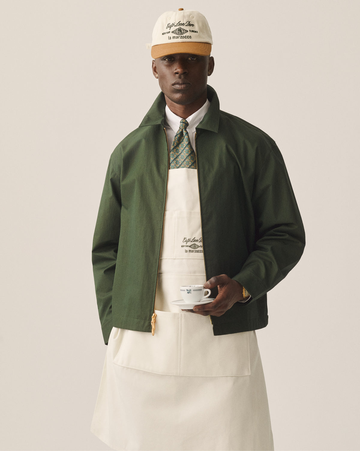 A man in a green jacket and apron wears the Café Leon Dore / La Marzocco Hat, holding a white cup and saucer.