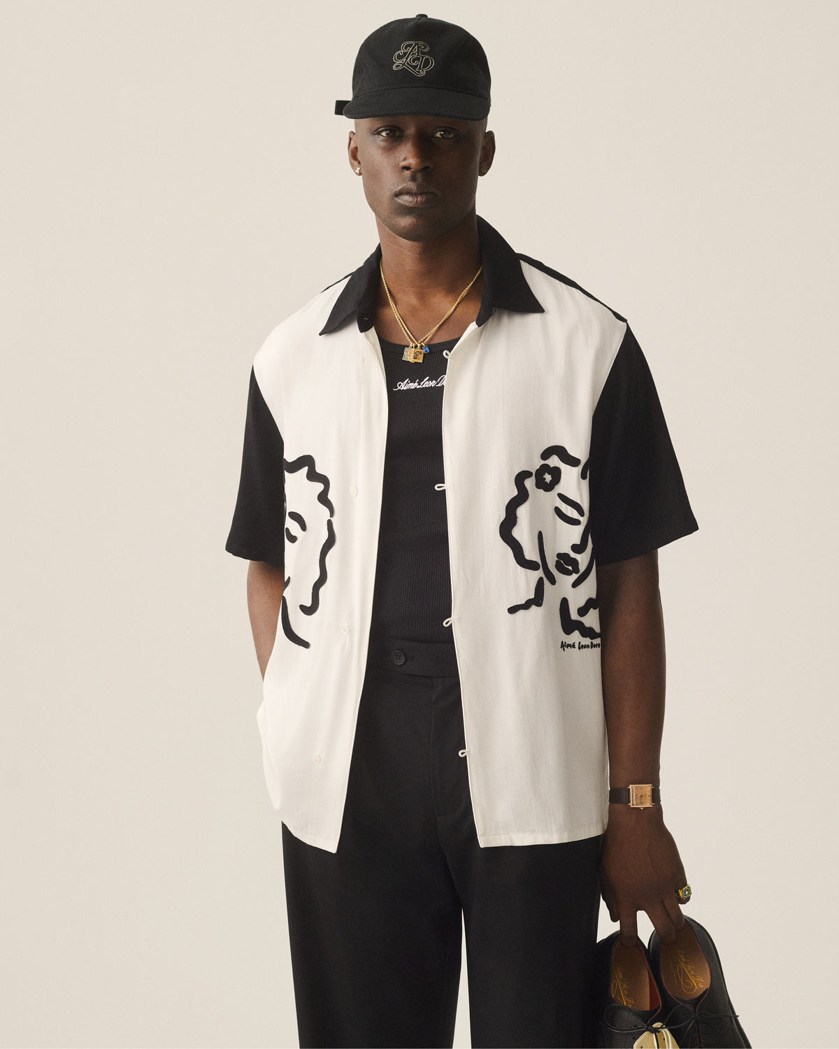 A man in an Embroidered Logo Tank Top, black pants, and cap holds shoes against a plain background.