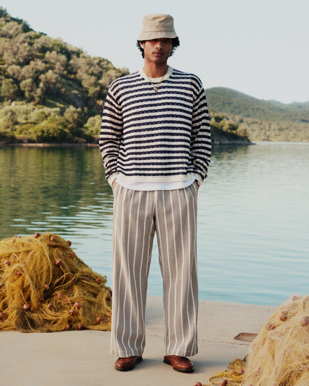 A person in a striped sweater and Wide Leg Shore Pant stands on a dock by the water with fishing nets and hills behind.
