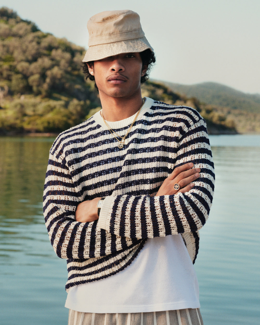 A man in a Skáfos Sweater and bucket hat stands by a lake with trees and hills in the background.