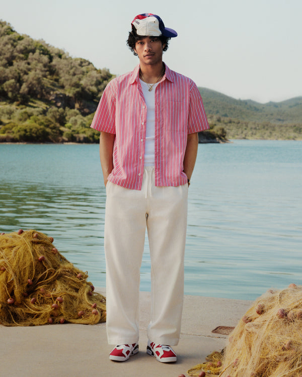 A man wearing Wide Leg Shore Pants and a red striped shirt stands by a lake with nets and green hills behind him.