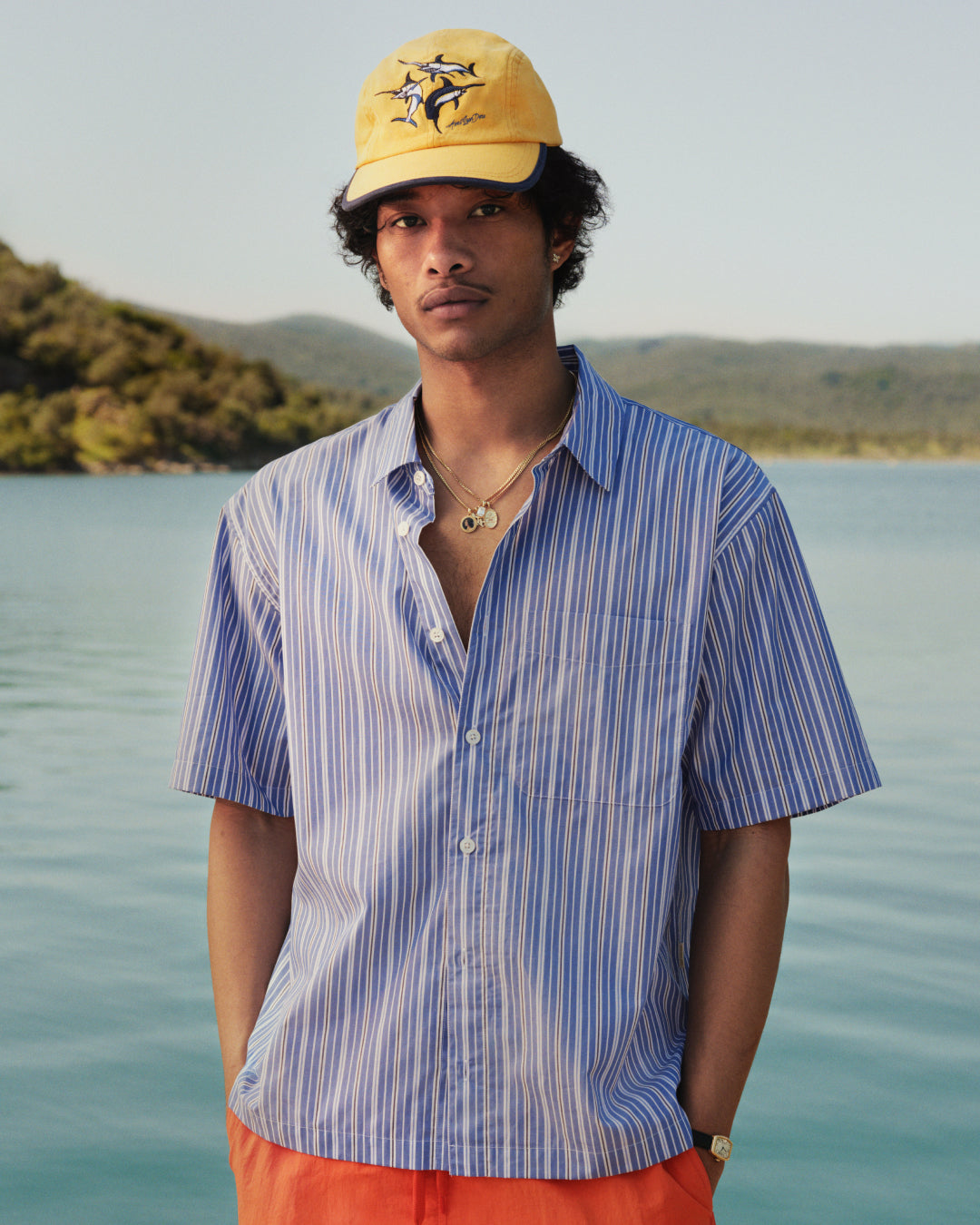 Man in a yellow cap, Yarn Dyed Striped Shirt, and orange shorts stands by a calm lake with hills in the background.