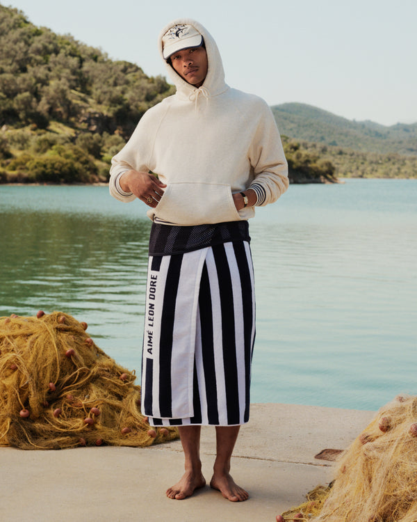 A man in a hoodie and Striped Beach Towel stands barefoot by a lake, with fishing nets and trees behind him.