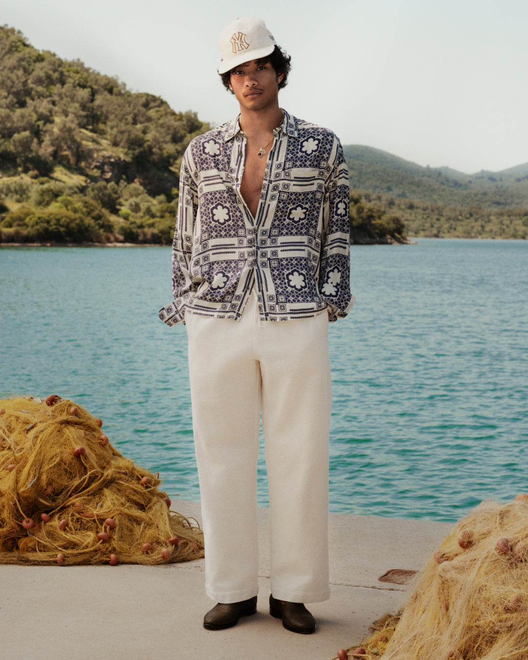 Man in light pants, Mosaic Button Down Shirt, and white cap stands on a dock by water with mountains and fishing nets.