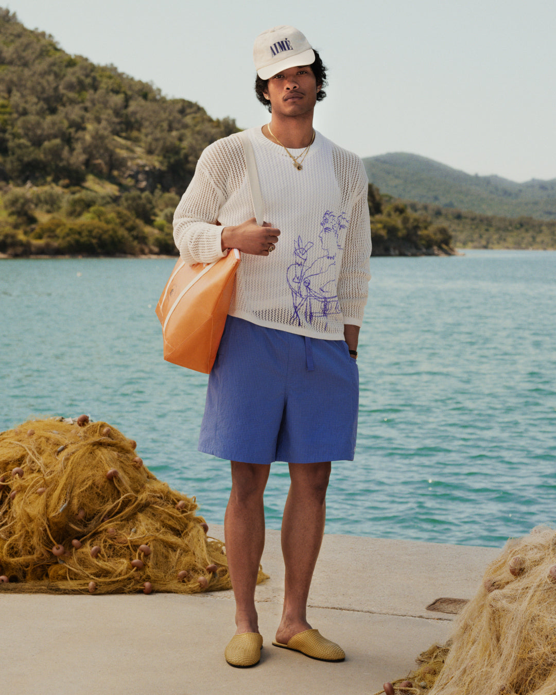 Man by a lake with mountains holds the orange Carryall Marina Tote, wearing a white sweater, blue shorts, and cap.