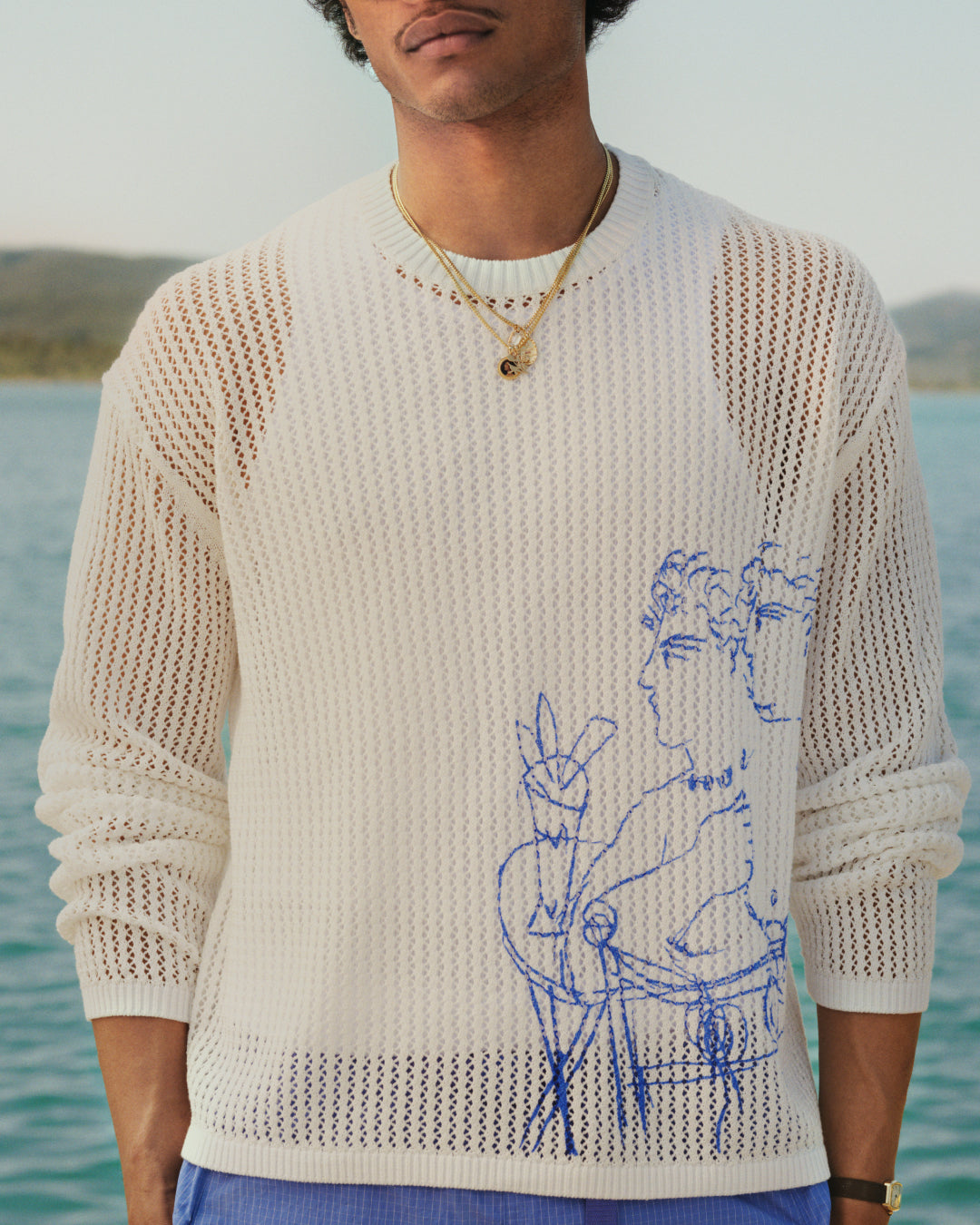 Someone in a Fassianos "Rendez - Vous" Crewneck Sweater with a blue figure sketch stands by the water.