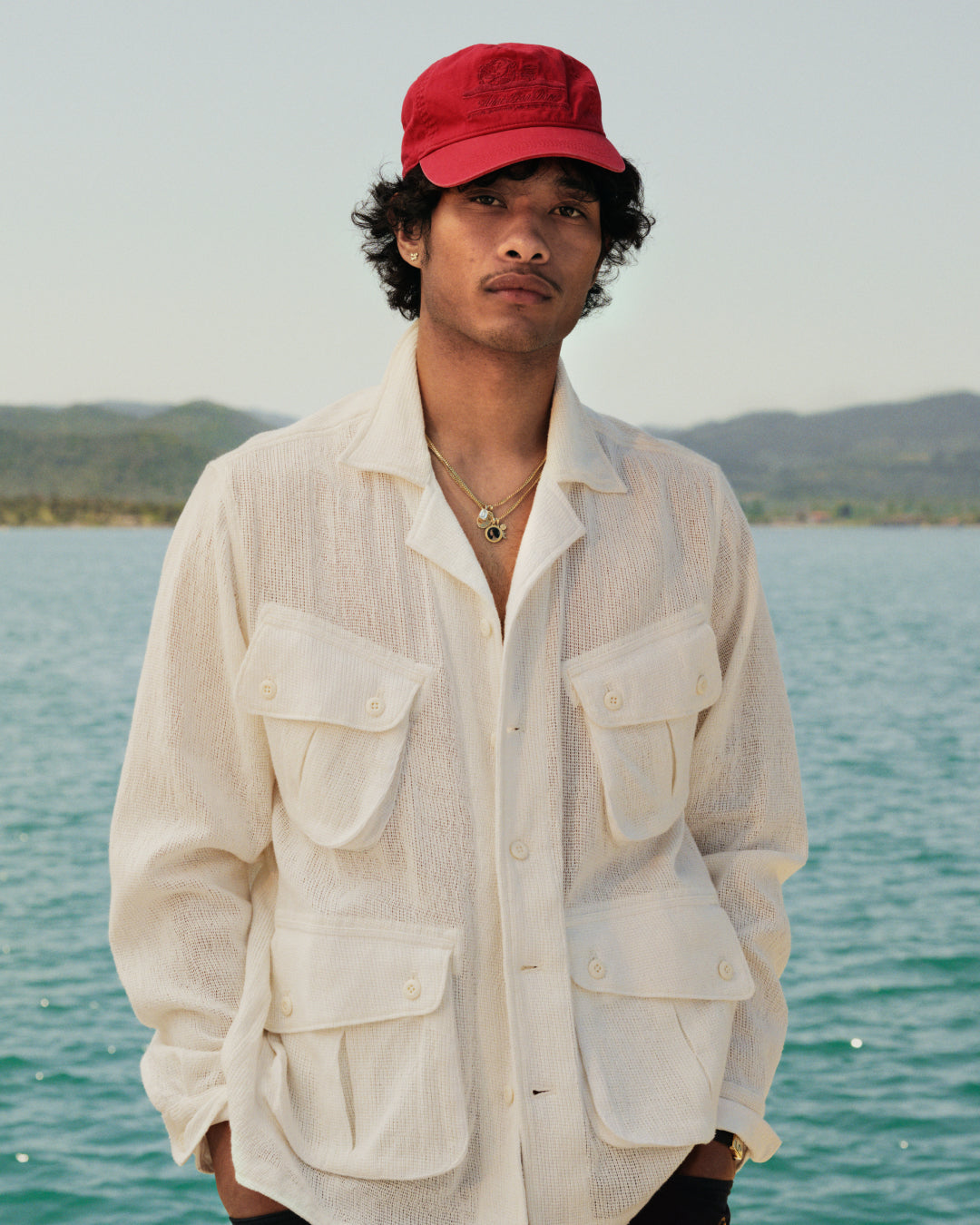 Man in a Mesh Jungle Shirt stands by water with mountains in the background.
