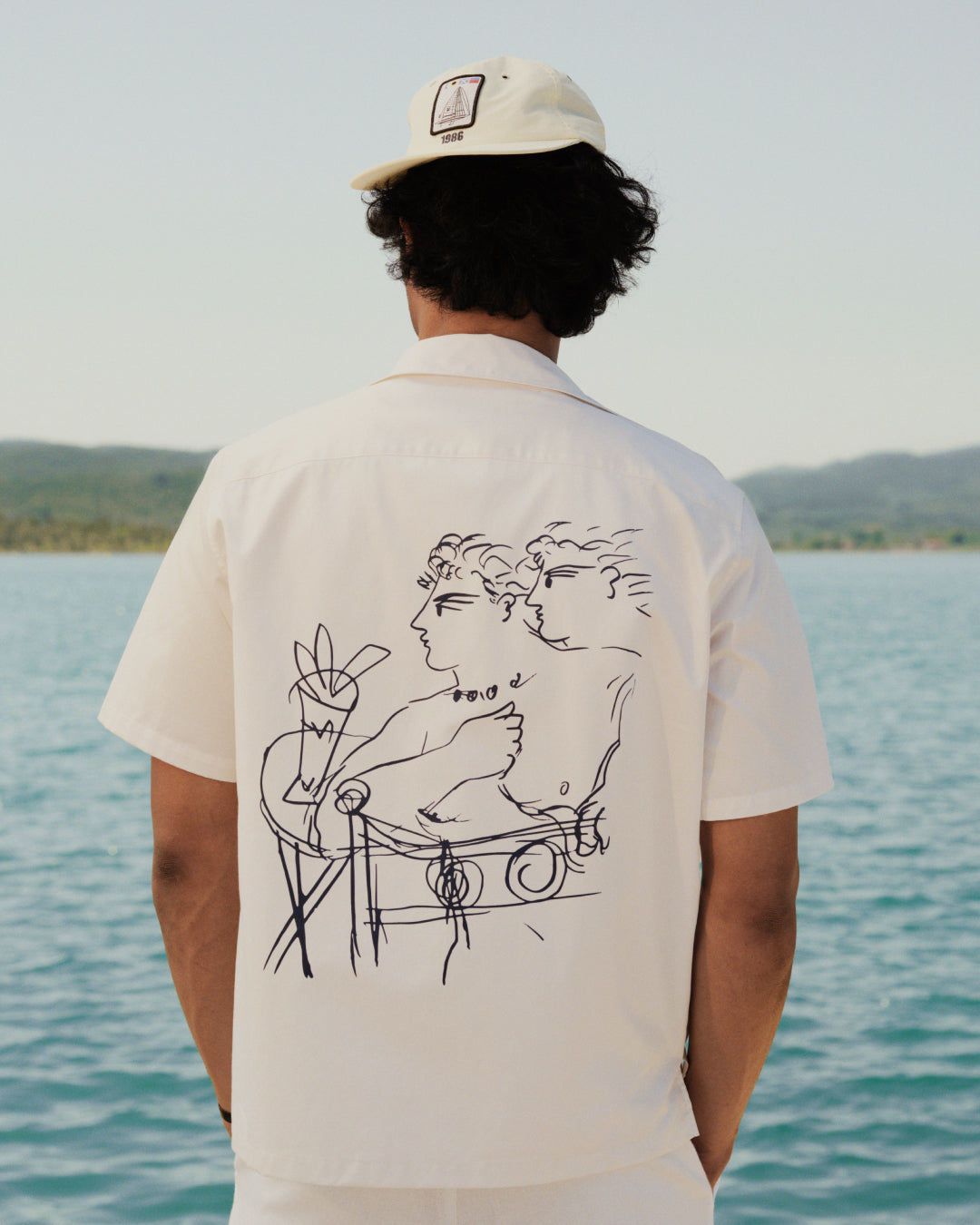 Someone wearing the Fassianos "Rendez - Vous" Camp Shirt stands by water, facing away from the camera.