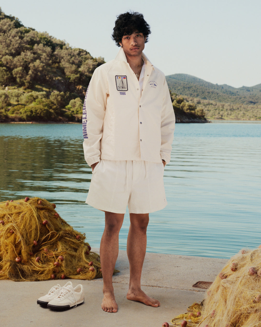 A man in a Marina Jacket stands barefoot on a dock by a lake, with shoes and fishing nets nearby, trees in the background.