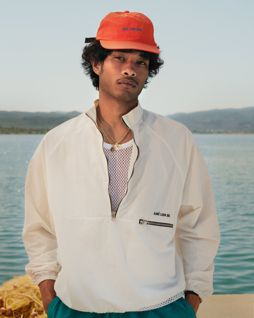 A man in a Nylon Packable Windbreaker and orange cap stands by water with hills in the background.