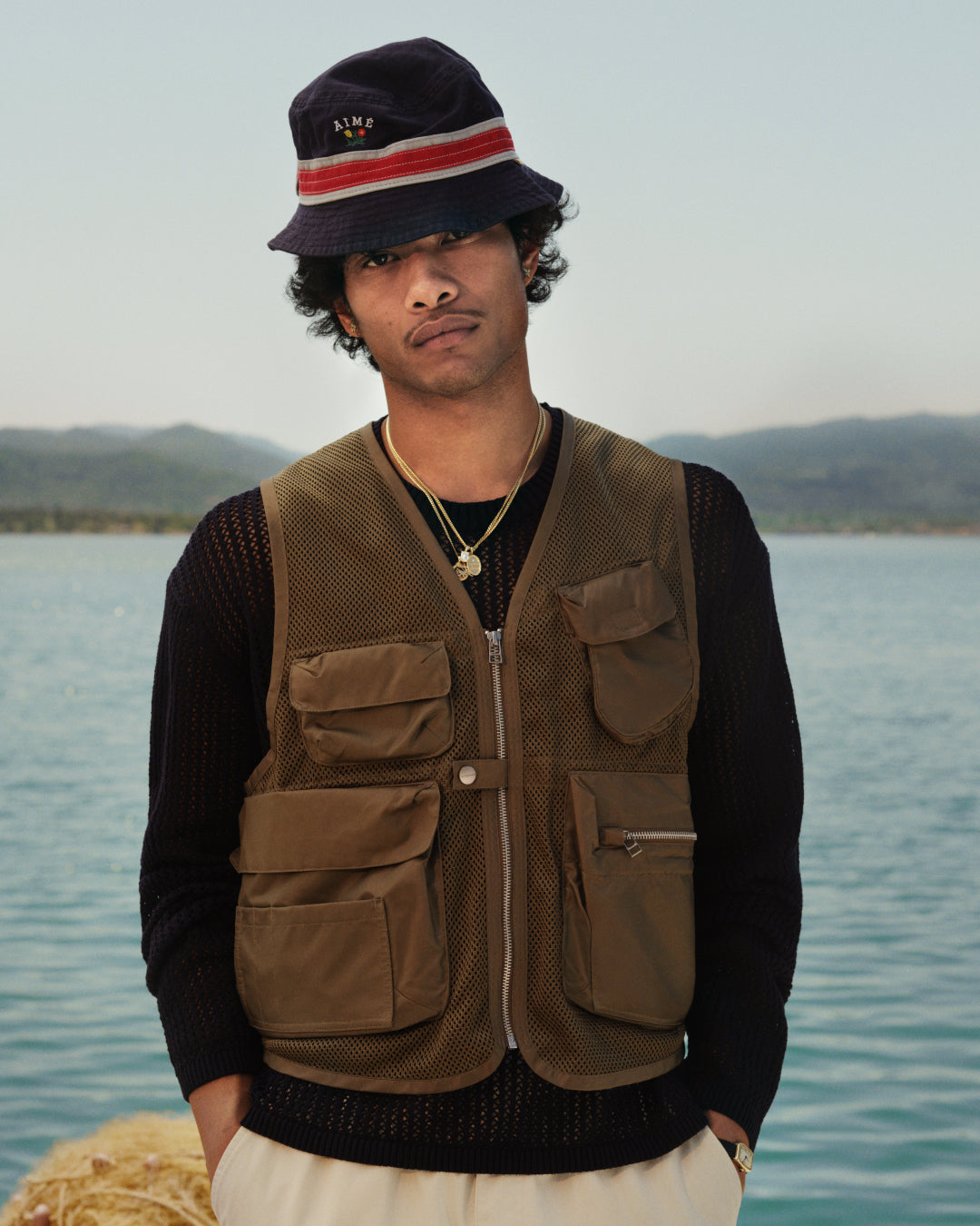 A man in a Cotton Eyelet Crewneck Sweater stands by a lake with mountains and trees in the background.