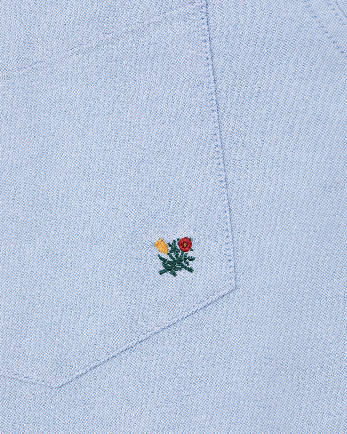 Light blue Crest Oxford Shirt with a pocket and a small yellow and red flower embroidered near the pocket's bottom.