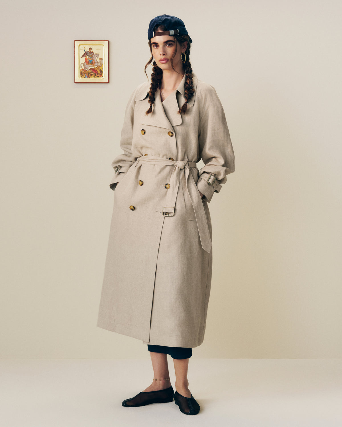Woman in a Linen Trench Coat, dark cap, and black shoes stands by framed art against a plain background.