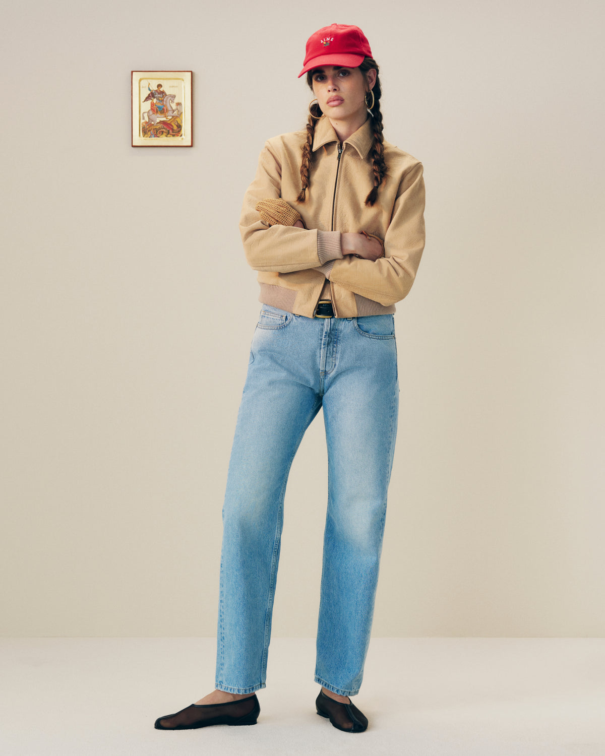 Woman in a tan jacket, light blue jeans, red cap, and Mesh Ballet Flats stands against a plain wall with framed art.
