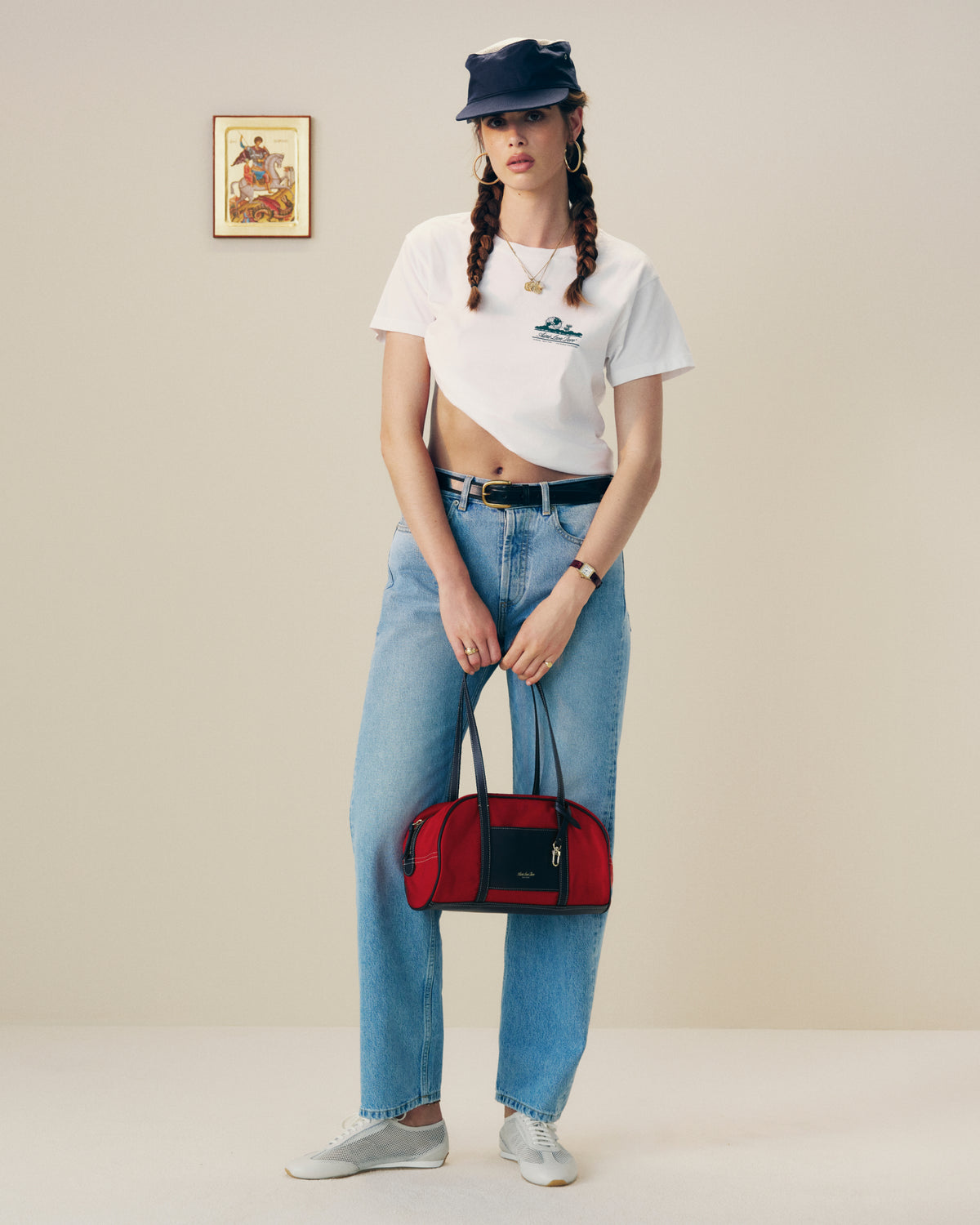 Woman in a white crop top, blue jeans, and black cap holds a red Mini Duffle Shoulder Bag in a minimal room.