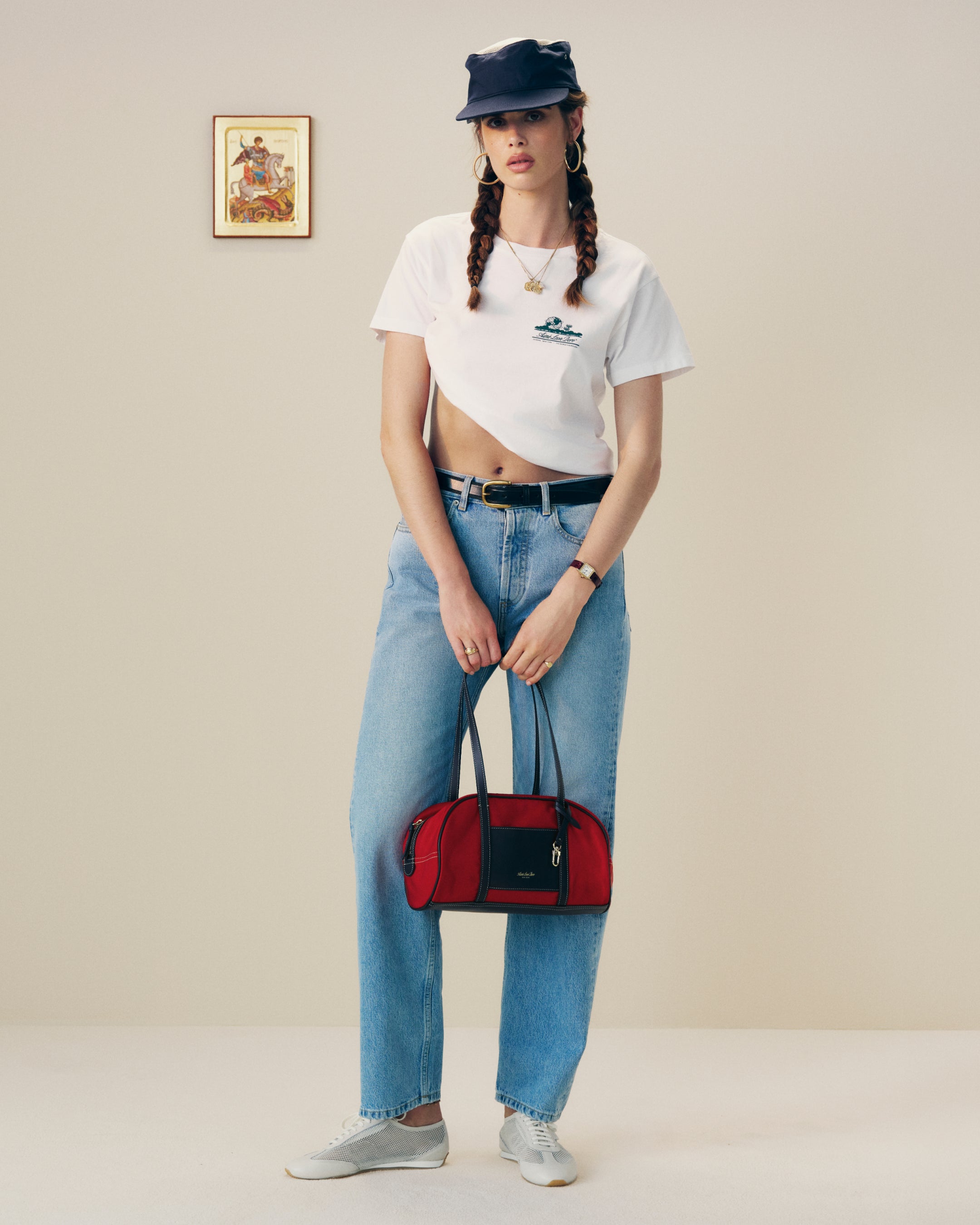 Woman in a white crop top, blue jeans, and black cap holds a red Mini Duffle Shoulder Bag in a minimal room.