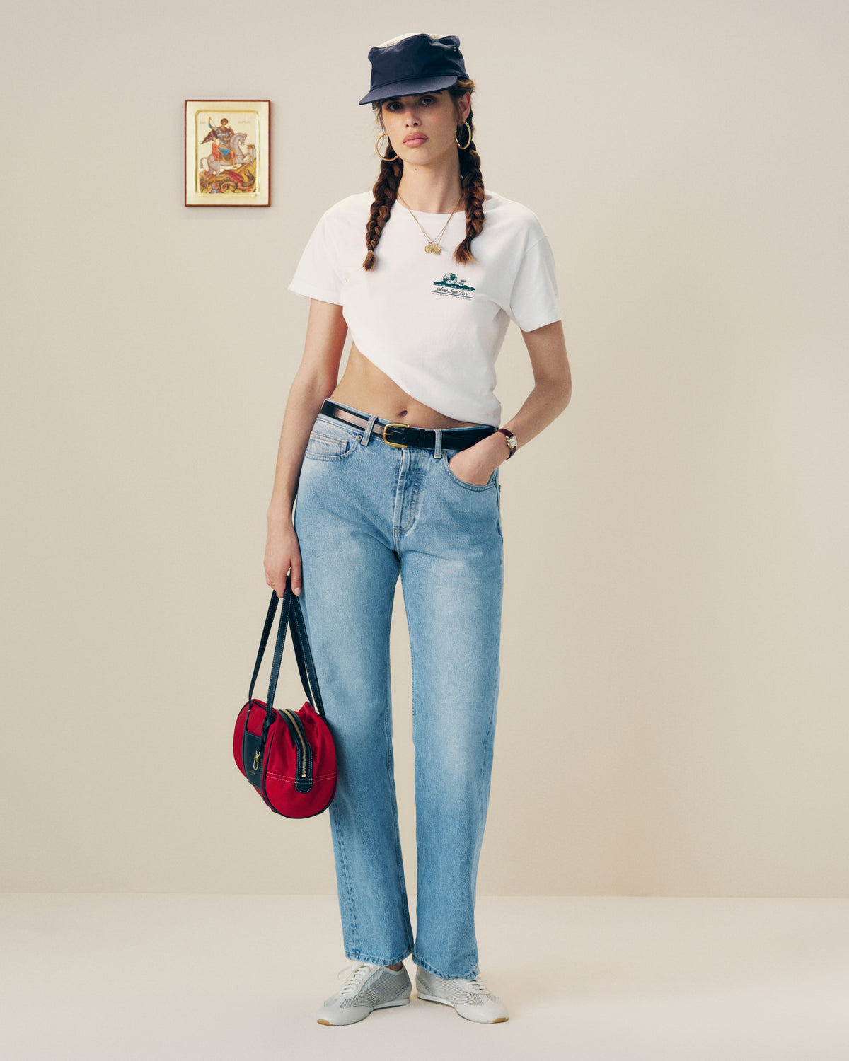 A woman in a Cropped Aloe-Blend Unisphere Tee, jeans, and cap holds a red bag in a minimalist art-filled room.