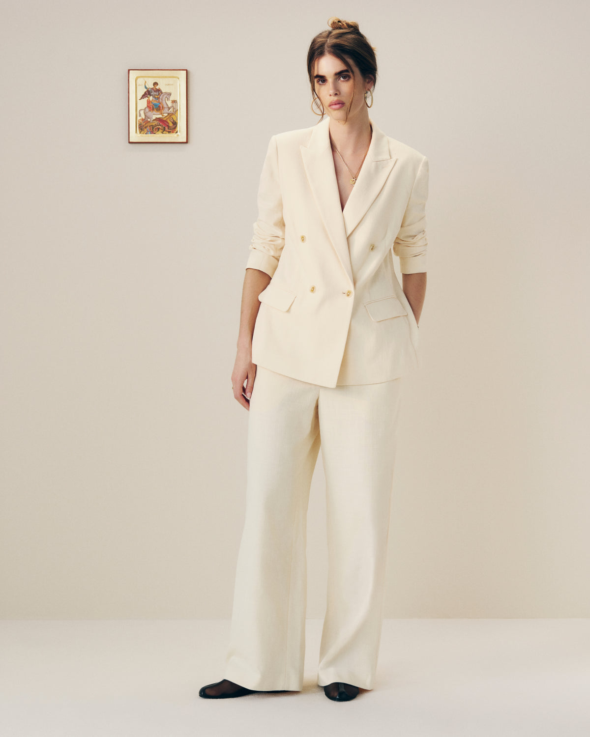 A woman in a Double-Breasted Unconstructed Blazer stands by a plain wall with art, wearing black shoes.