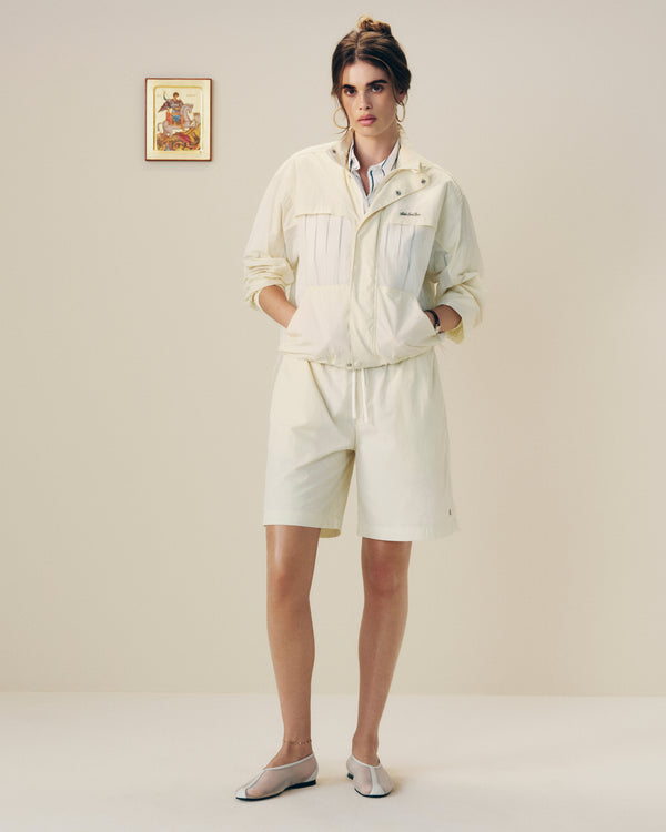 A person wears a Nylon Track Jacket and shorts indoors, hands in pockets, standing before a plain wall with a small picture.
