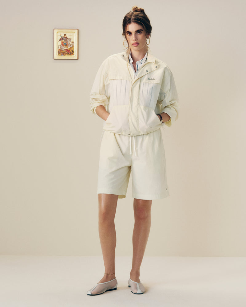 A person wears a Nylon Track Jacket and shorts indoors, hands in pockets, standing before a plain wall with a small picture.