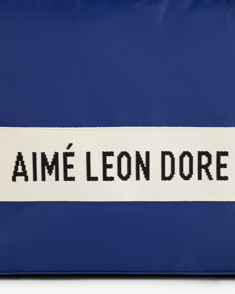 Blue nylon duffle bag with a white center stripe and black AIMÉ LEON DORE text.