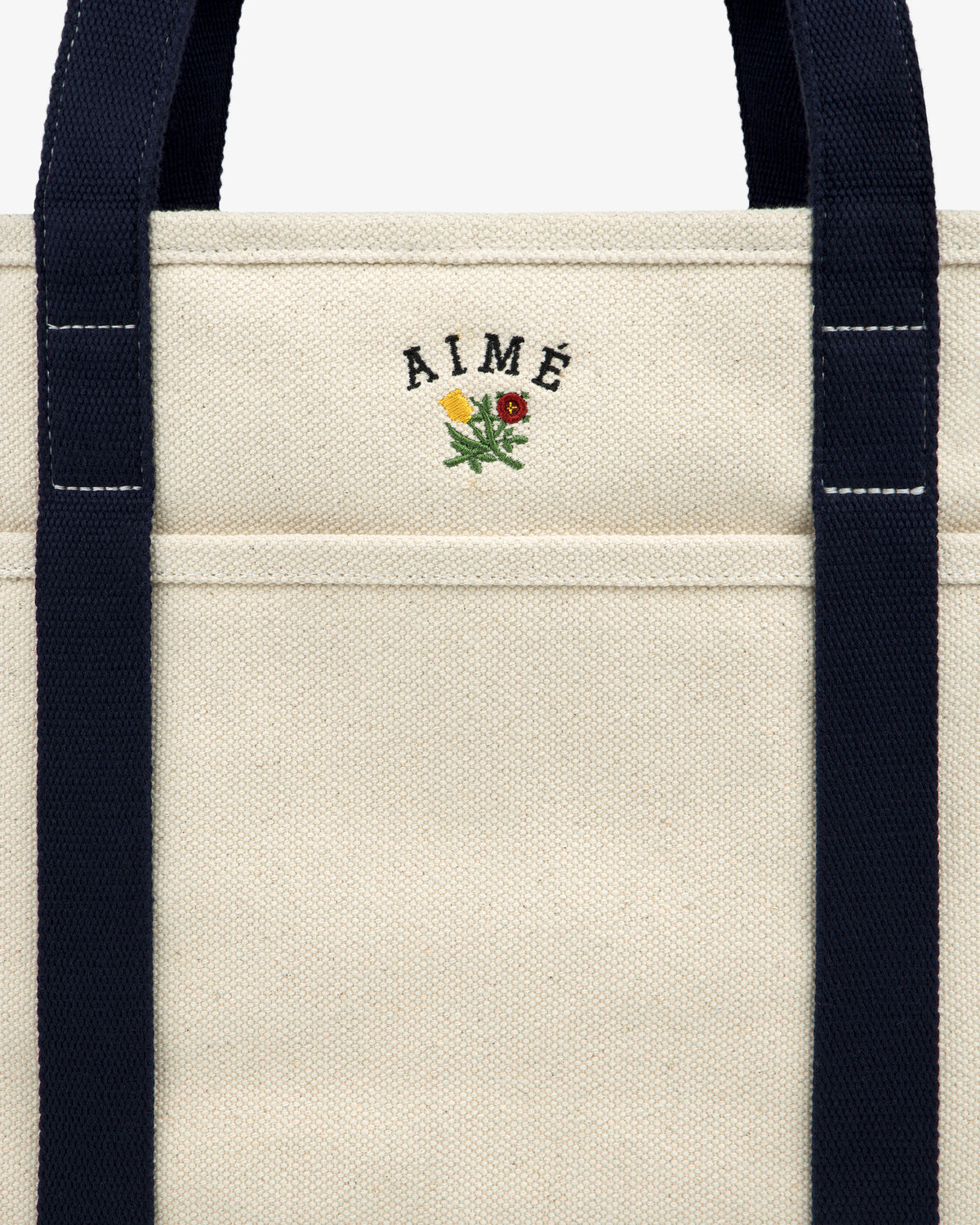 Small Crest Contrast Tote Bag in beige canvas with navy straps, AIMÉ embroidery, and a floral crest above the pocket.