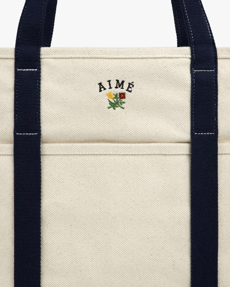 Close-up of the Large Crest Contrast Tote Bag in beige canvas with AIMÉ, floral embroidery, and navy blue handles.