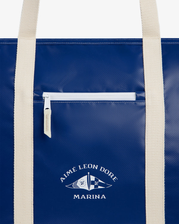 Carryall Marina Tote: blue bag with cream handles, front zipper pocket, and Aimé Leon Dore flag logo.