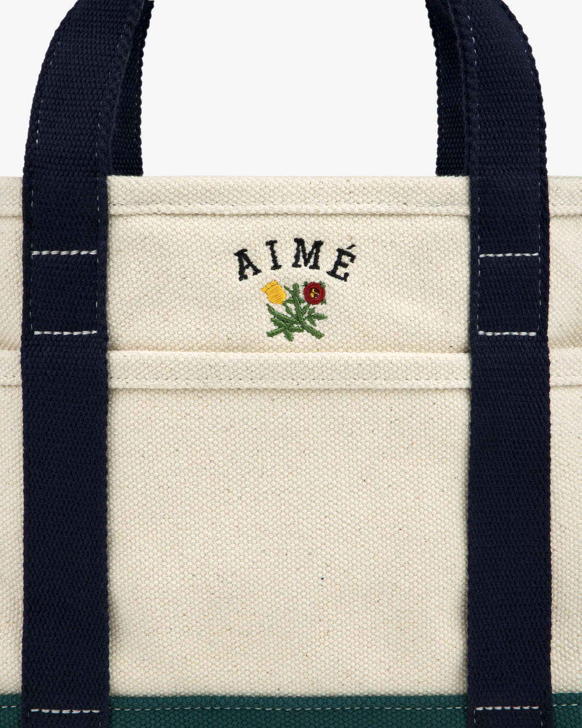 Close-up of the Mini Crest Contrast Tote Bag: beige canvas with navy handles, AIMÉ text, and a yellow-red flower.
