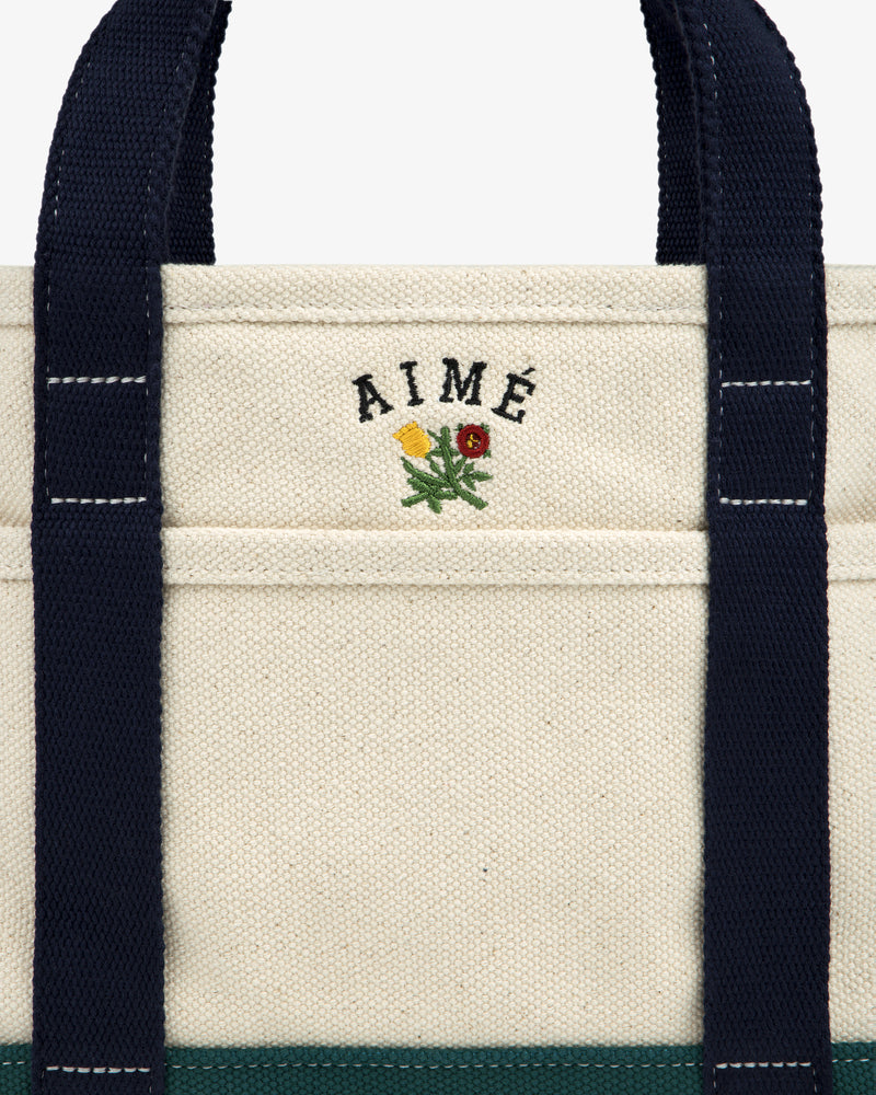 Close-up of the Mini Crest Contrast Tote Bag: beige canvas with navy handles, AIMÉ text, and a yellow-red flower.