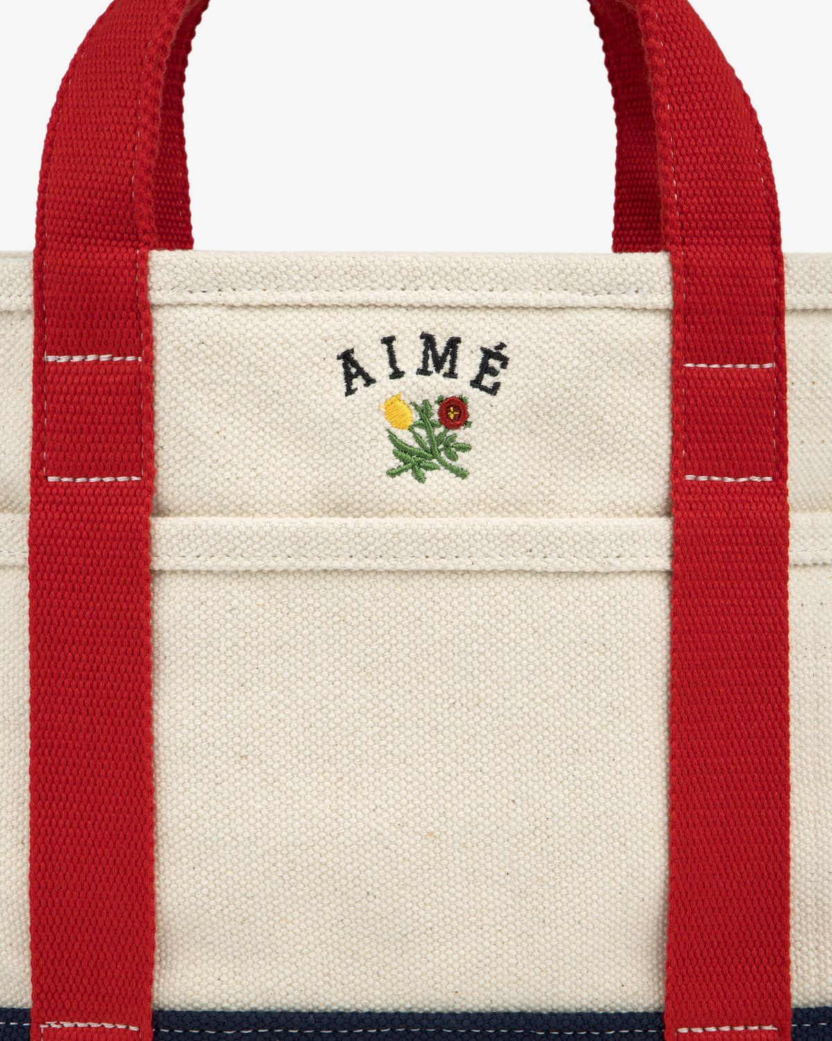 Close-up of the Mini Crest Contrast Tote Bag—cream canvas with red handles, AIMÉ text, and a flower above the pocket.