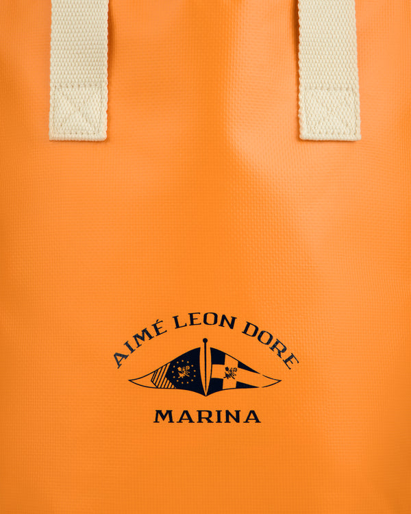 Close-up of the Marina Bottle Tote—an orange bag with cream handles and a navy AIMÉ LEON DORE MARINA logo and flags.