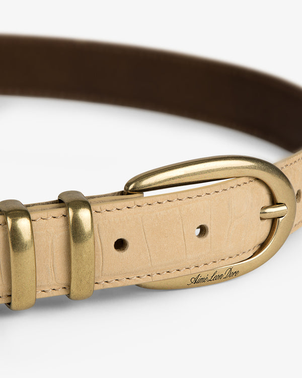 Close-up of a Croc-Embossed Nubuck Belt in beige with a gold buckle and Ami. Son Dore engraved near the buckle.