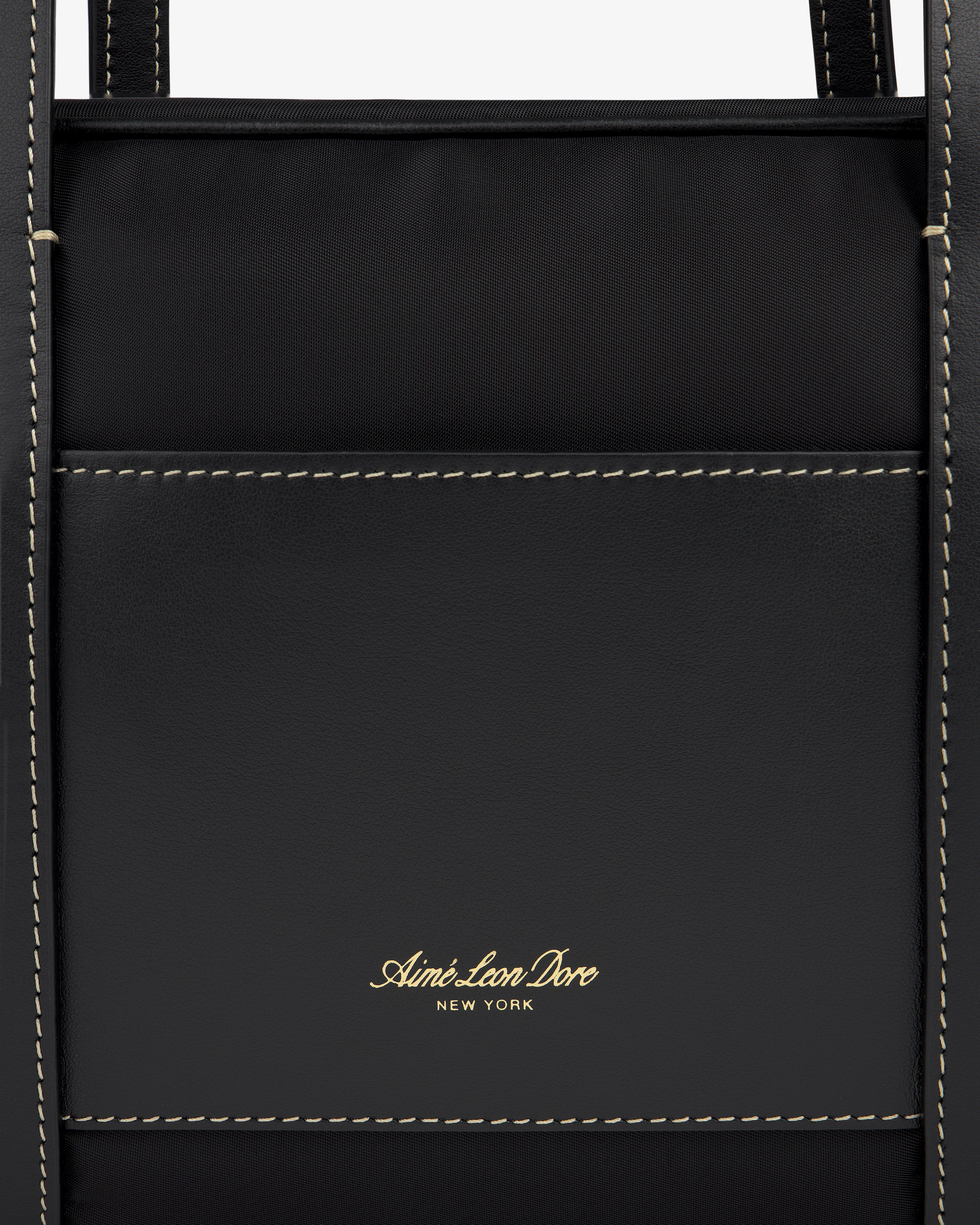 Close-up of a black Mini Duffle Shoulder Bag by Aimé Leon Dore with white stitching and gold logo on front pocket.