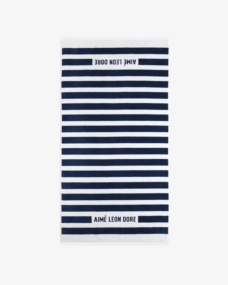 The Striped Beach Towel is white with navy blue horizontal stripes and AIMÉ LEON DORE text on both ends.