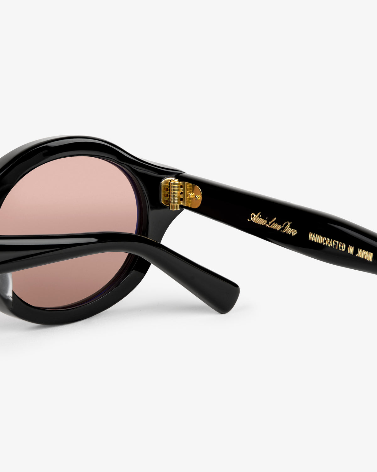 Close-up of Onásis Sunglasses: black frame, pink lenses, and gold "Handcrafted in Japan" text on the arm.