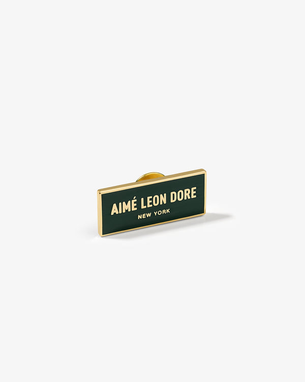 Graphic Pin with gold trim features AIMÉ LEON DORE NEW YORK text on a dark green rectangular background.