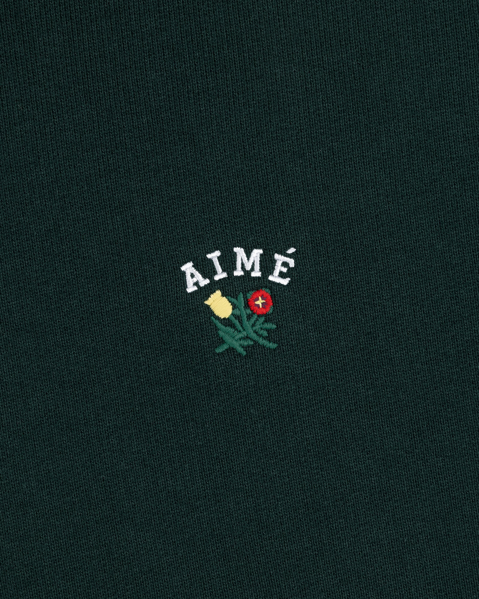 The Crest Hoodie features "AIMÉ" embroidered above yellow and red flowers on dark green fabric.